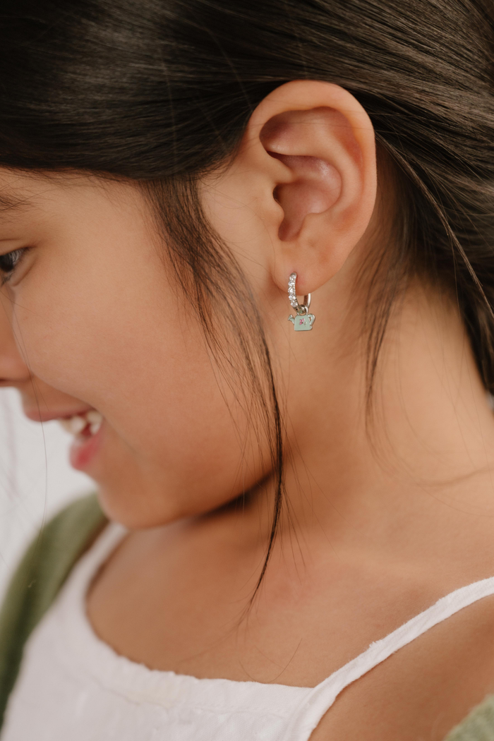 A close up of a right-facing watering can shaped charm with mint green enamel and titanium metal hanging from a hoop earring.