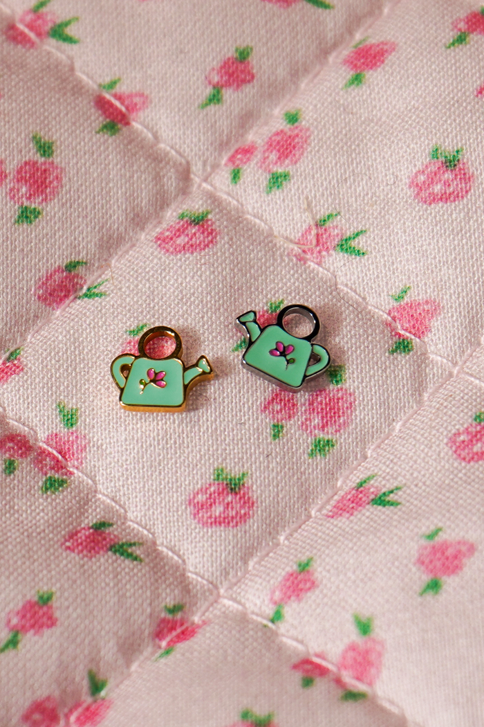 A close up of 2 watering can shaped hoop charms with mint green enamel and titanium metal on a pink floral background