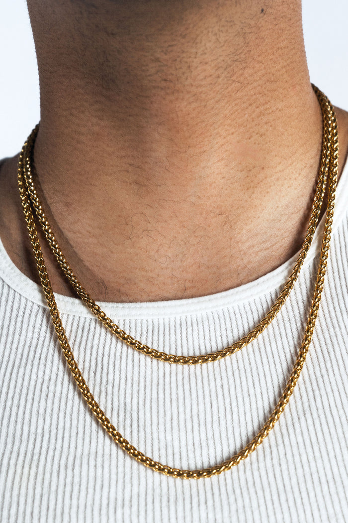 A close-up of a person's neck and upper chest wearing a double-layered gold chain necklace over a white ribbed tank top.