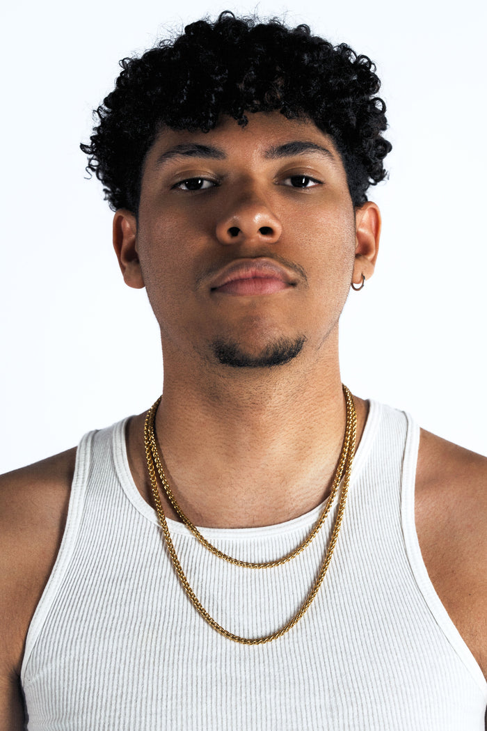 A man with curly black hair wearing a white ribbed tank top and two gold chain necklaces, photographed against a plain white studio background.