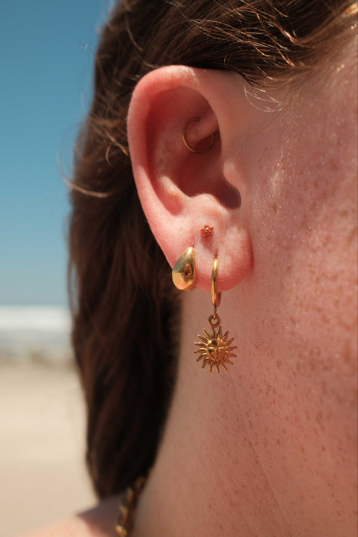 Close-up of an ear wearing gold hoop earrings with a sun design, with blurred background.