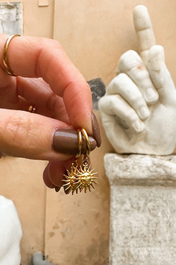 Gold earrings with a textured sun design held by a hand in front of a marble statue.